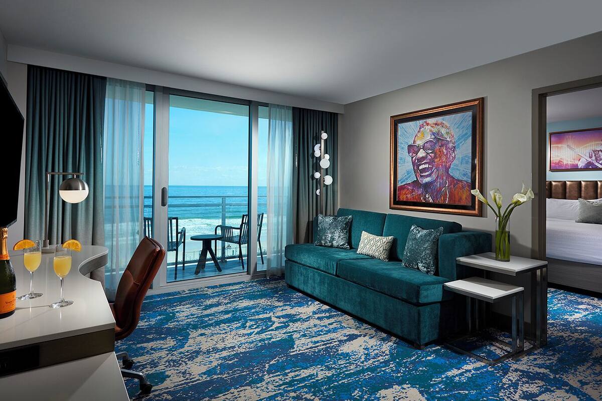 A hotel room with a large window offering a view of the ocean, a teal sofa, a small table with a vase of flowers, and a bed with a TV on the wall.