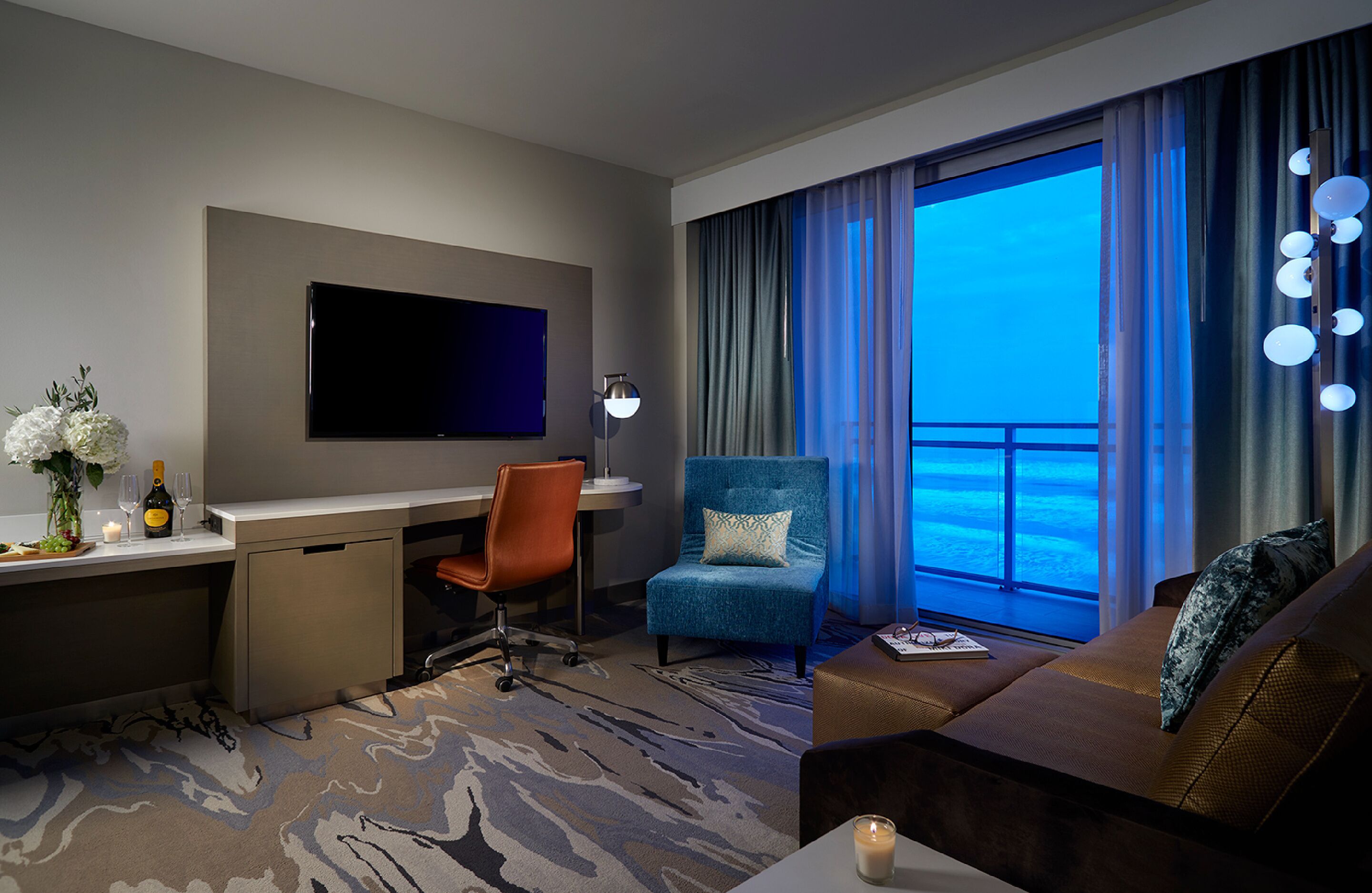 A hotel room with a large TV, a desk with a chair, a blue armchair, a sofa, and a view of the ocean.