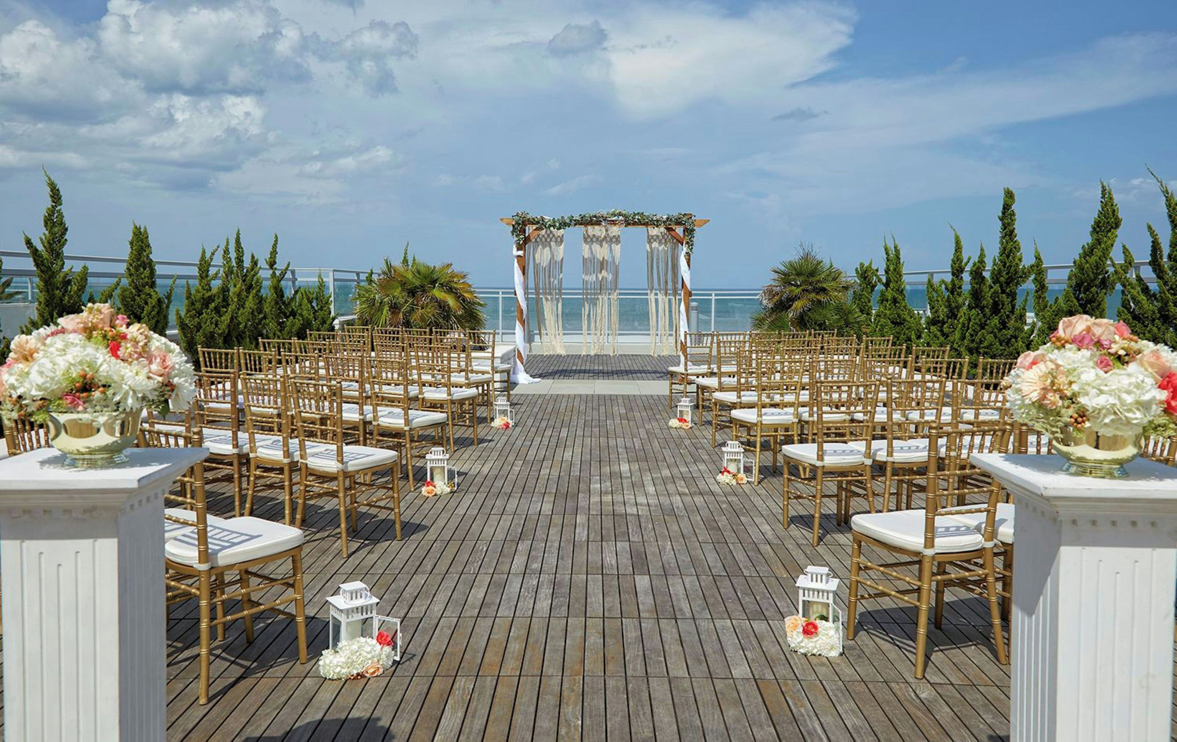 An outdoor wedding ceremony setup with a wooden deck, white chairs, and a floral arch.