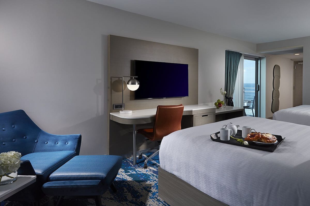 A hotel room with a bed, a desk, a chair, a TV, and a view of the sea.