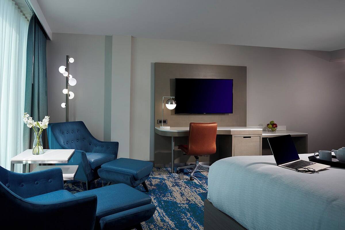 A hotel room with a bed, two blue armchairs, a desk with a laptop, a TV, and a small table with a vase of flowers.