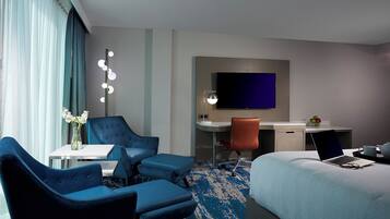 A hotel room with a bed, two blue armchairs, a desk with a laptop, a TV, and a small table with a vase of flowers.