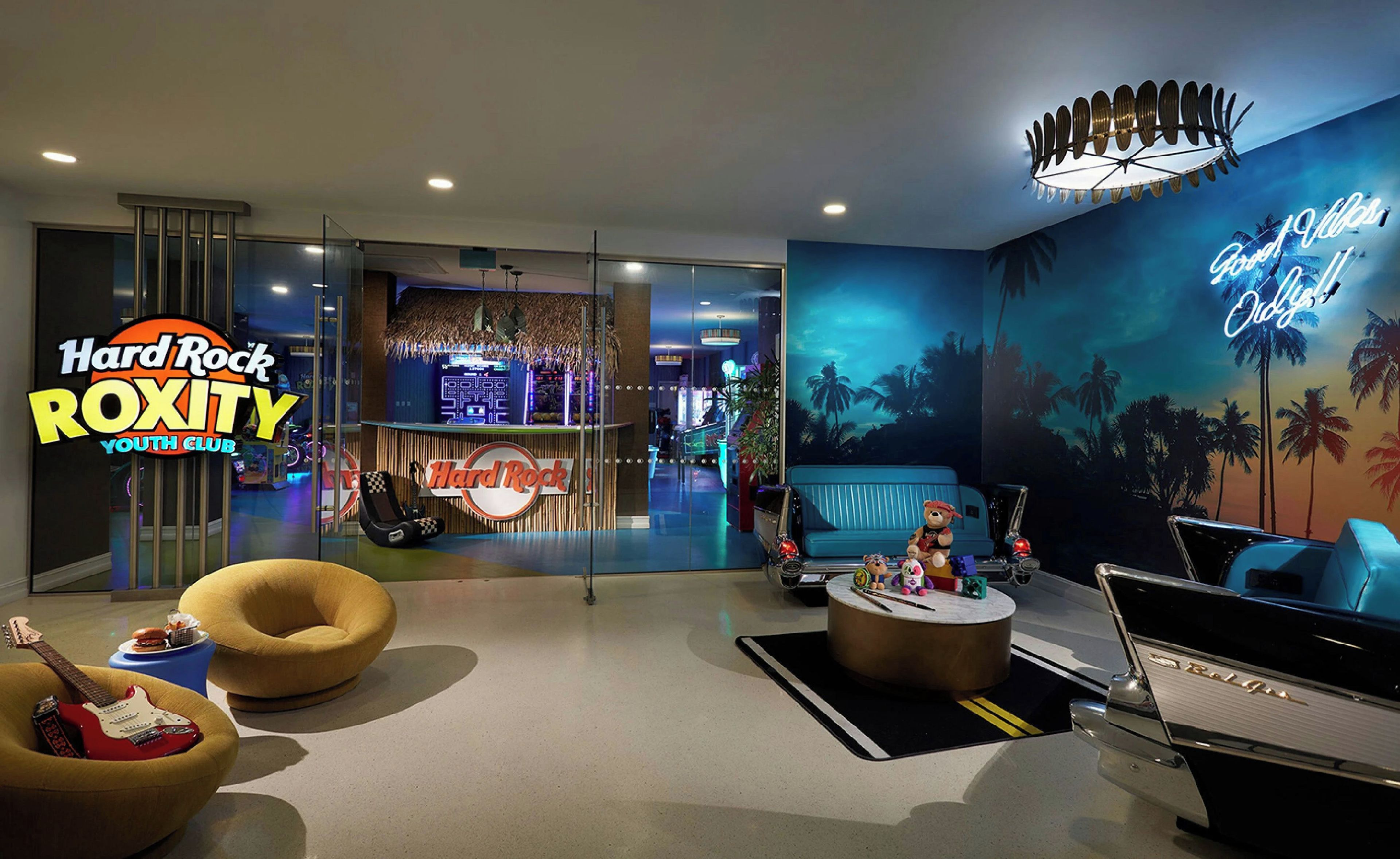A Hard Rock Roxy Youth Club with a tropical mural, a neon sign, a vintage car, and a guitar-shaped chair.