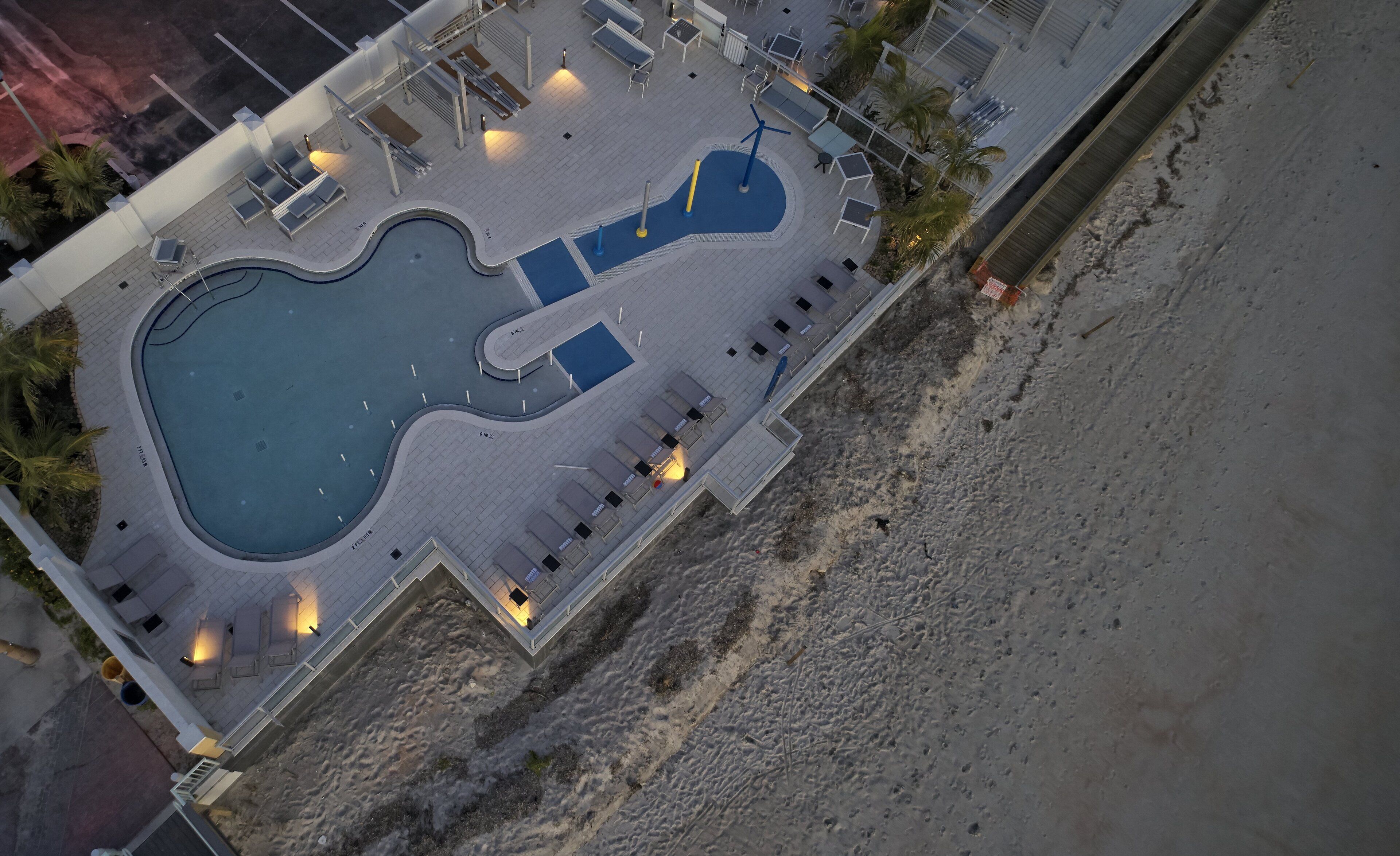An aerial view of a resort with a large swimming pool, sun loungers, and a beach area.