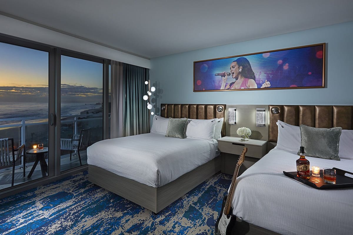 A hotel room with a large balcony, a bed with white linens, a bedside table with a bottle and glass, and a painting on the wall.