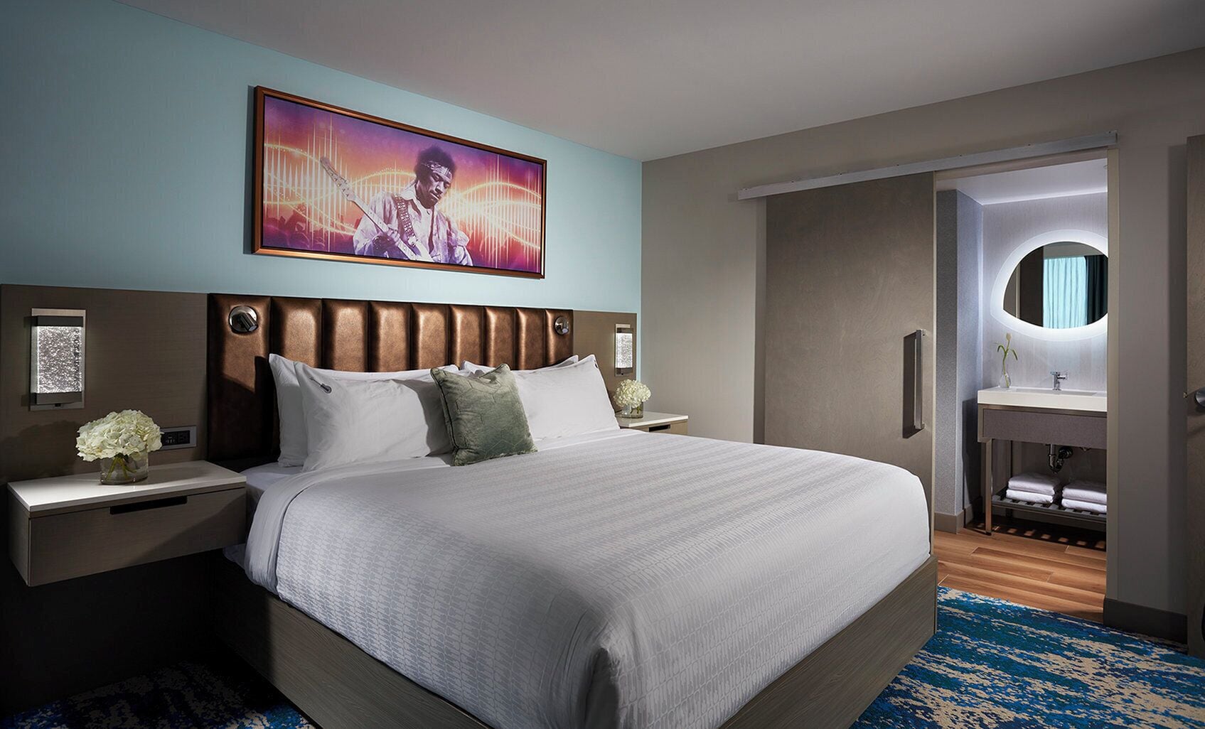 A hotel room with a large bed, a headboard with a light wood finish, a wall-mounted TV, a bedside table with a vase of flowers, and a bathroom visible through an open door.