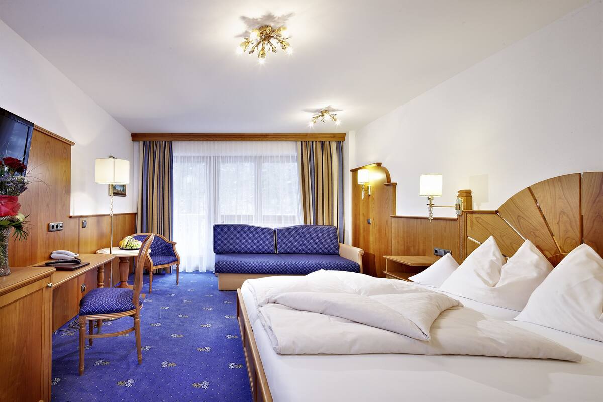 A hotel room with a bed, a desk, a chair, a sofa, and a large window with curtains.