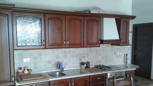 A kitchen with wooden cabinets, a tiled backsplash, a gas stove, and a dining table with a fruit bowl. - I Tufi (Colonnella)