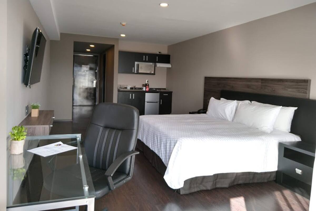 A modern hotel room with a large bed, a desk with a chair, a flat-screen TV, a kitchenette, and a dark wood headboard.