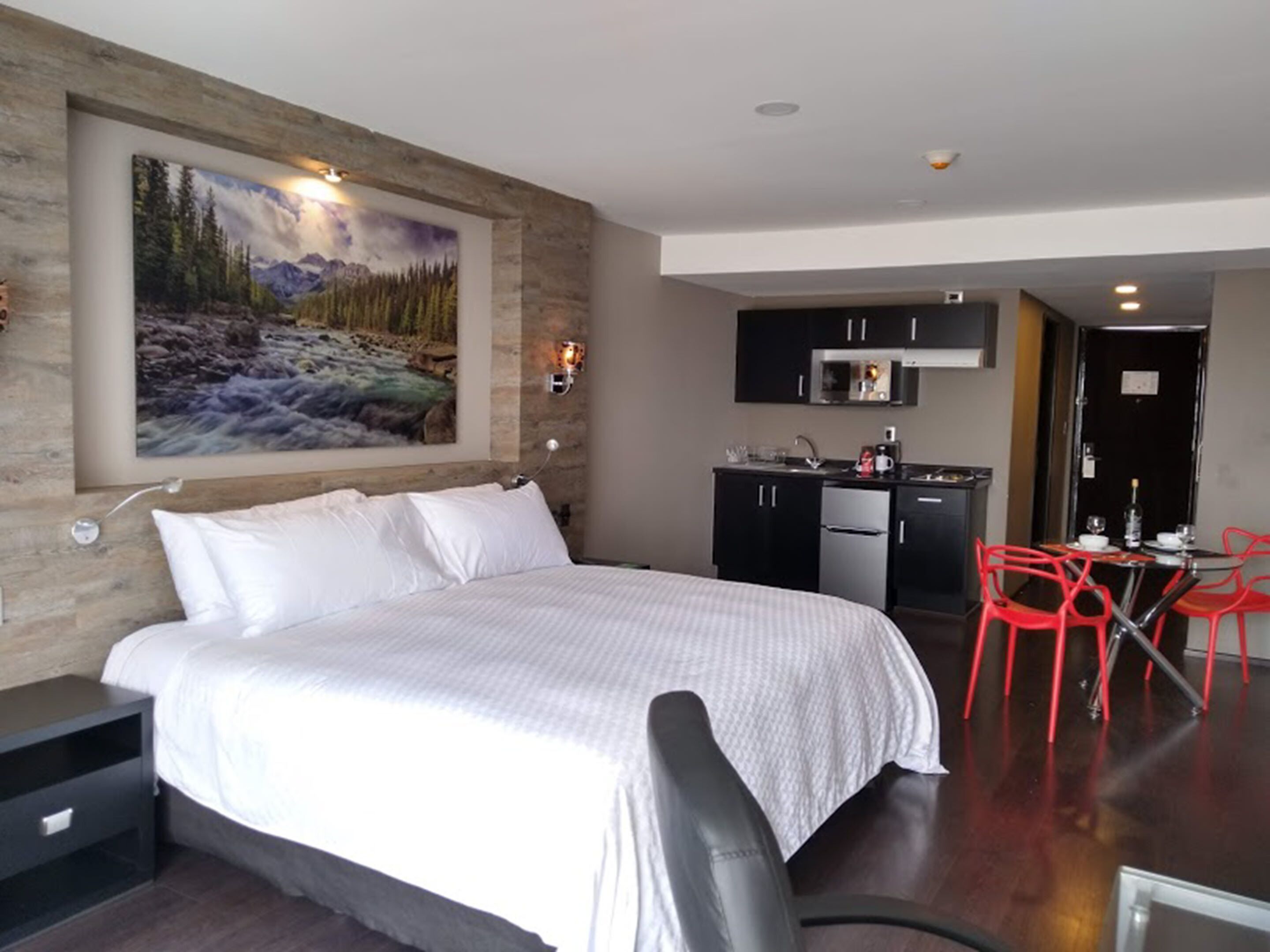 Una habitación de hotel con cama, un cuadro de un paisaje montañoso, una kitchenette y sillas rojas.