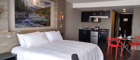 A hotel room with a bed, a painting of a mountain landscape, a kitchenette, and red chairs.