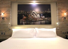 Una habitación de hotel con una cama grande, ropa de cama blanca y un cuadro de la ciudad que muestra la Casa de la Ópera y el Puente de Puerto de Sydney.