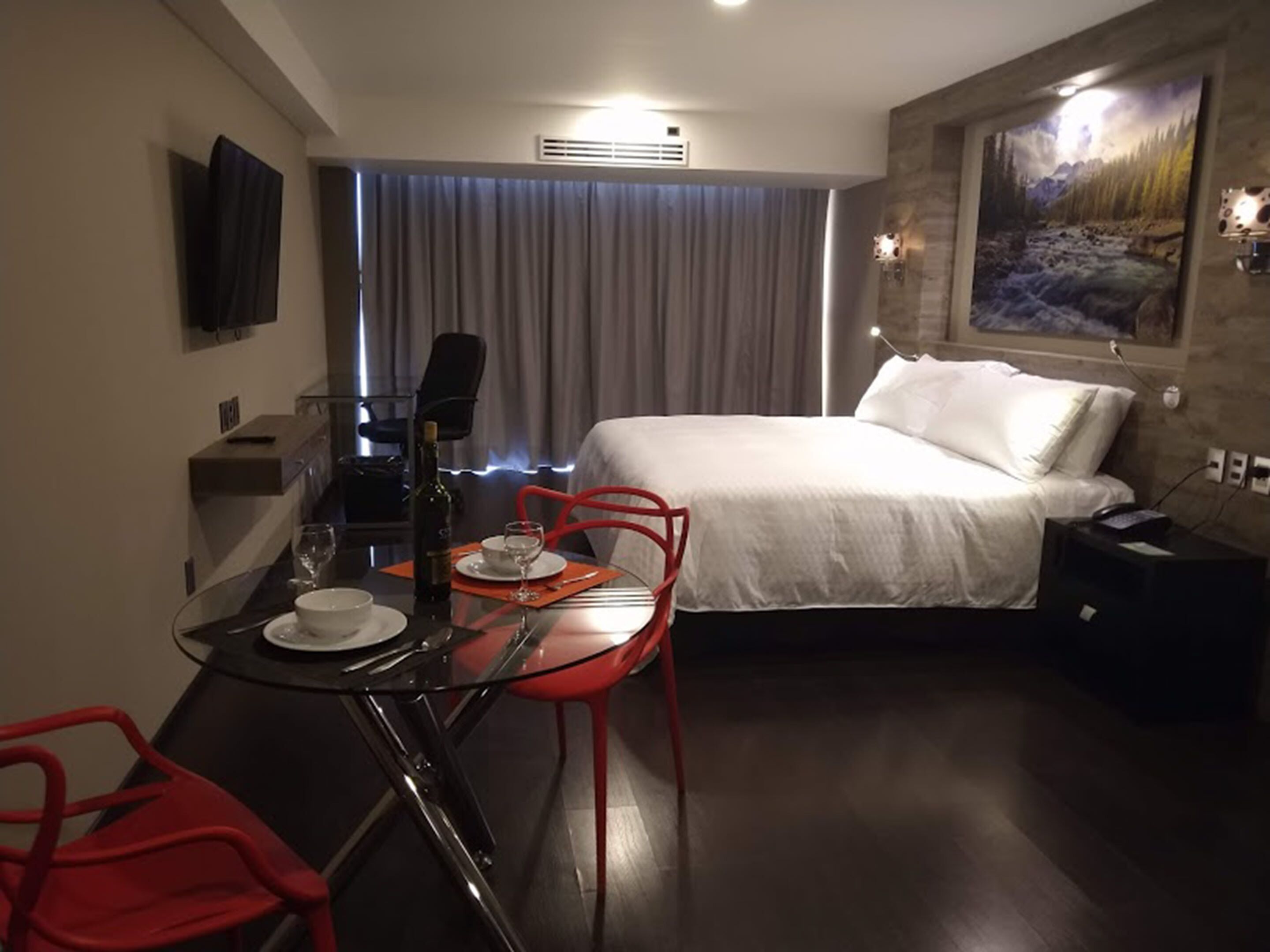 Habitación de hotel con cama, una mesa de comedor con sillas, un televisor montado en la pared y un cuadro colgado.