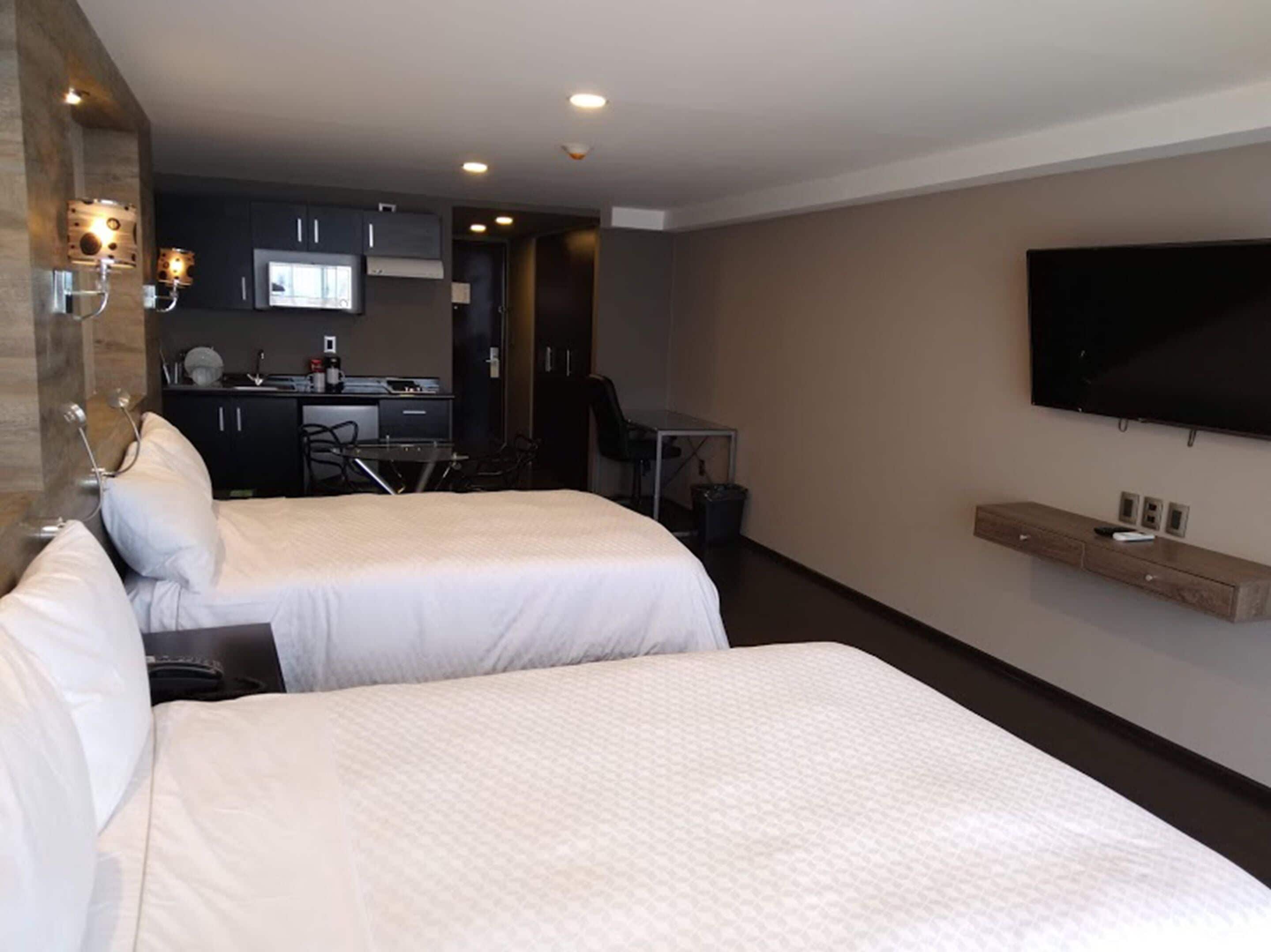 A hotel room with two beds, a TV mounted on the wall, a desk with a chair, and a kitchenette in the background.