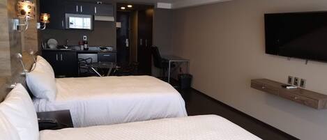 A hotel room with two beds, a TV mounted on the wall, a desk with a chair, and a kitchenette in the background.