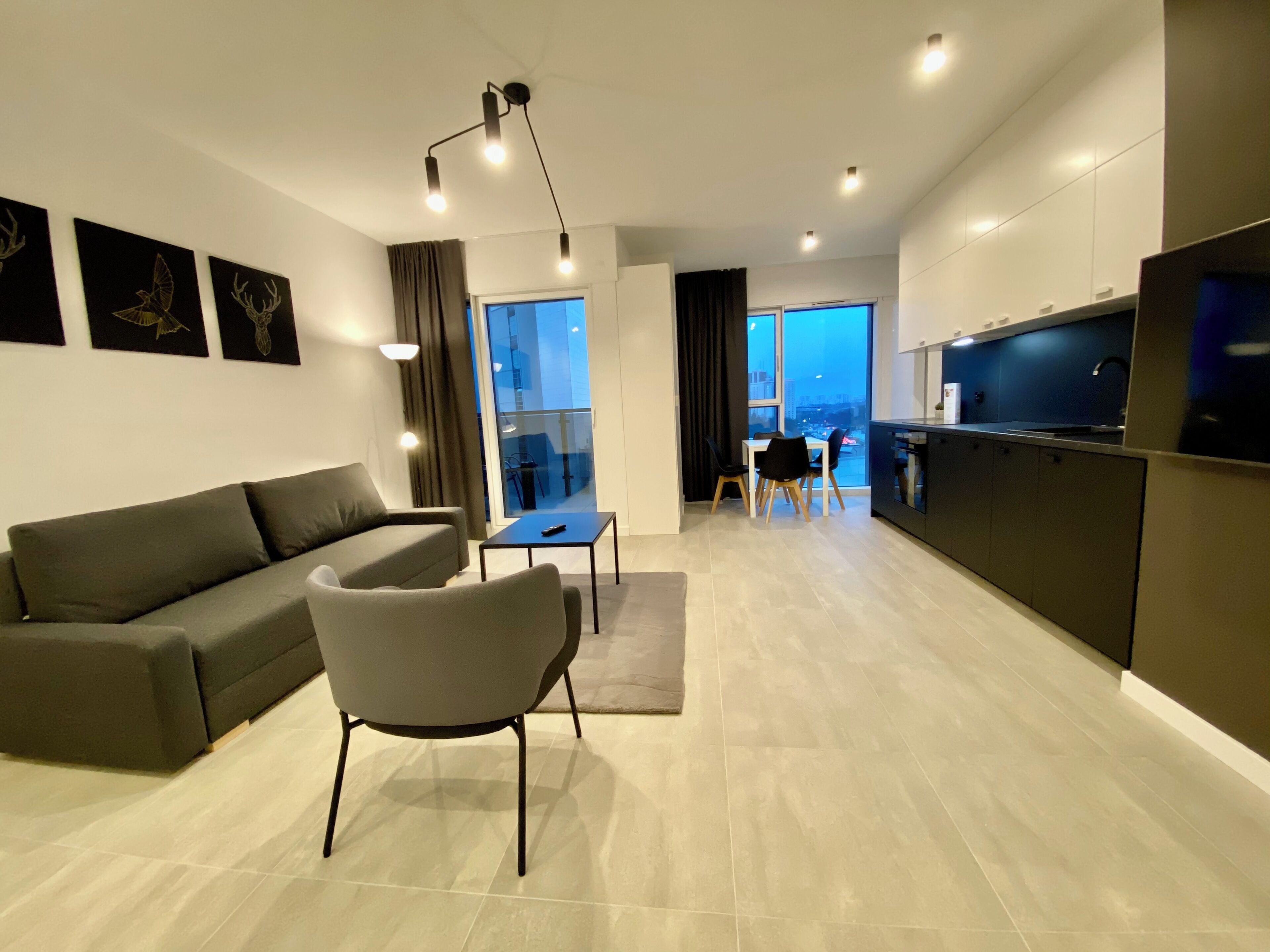 A modern living room with a sofa, a coffee table, and a TV. There is a dining area with chairs and a table. The room has a large window and a view of the city.