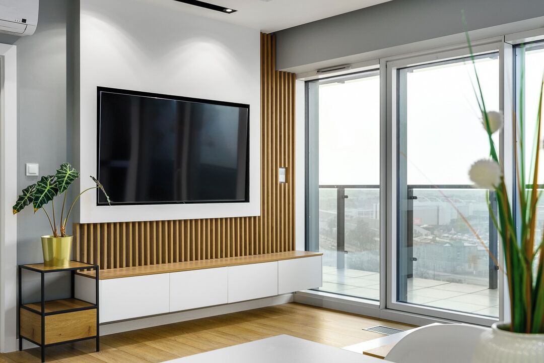 A modern living room with a large flat-screen TV mounted on the wall, a wooden accent wall, a sliding glass door leading to a balcony, and a small side table with a plant.