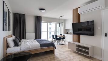 A modern hotel room with a bed, a TV mounted on the wall, a dining area with chairs, and a large window.