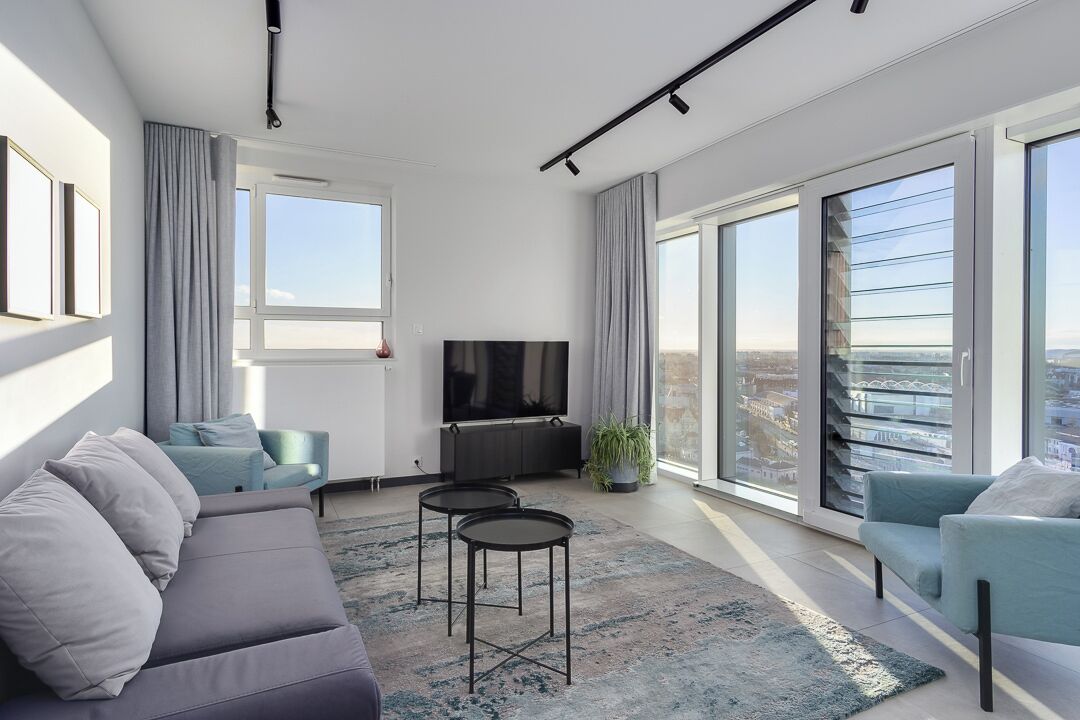A modern living room with a sofa, two armchairs, a TV, and a large window with a city view.