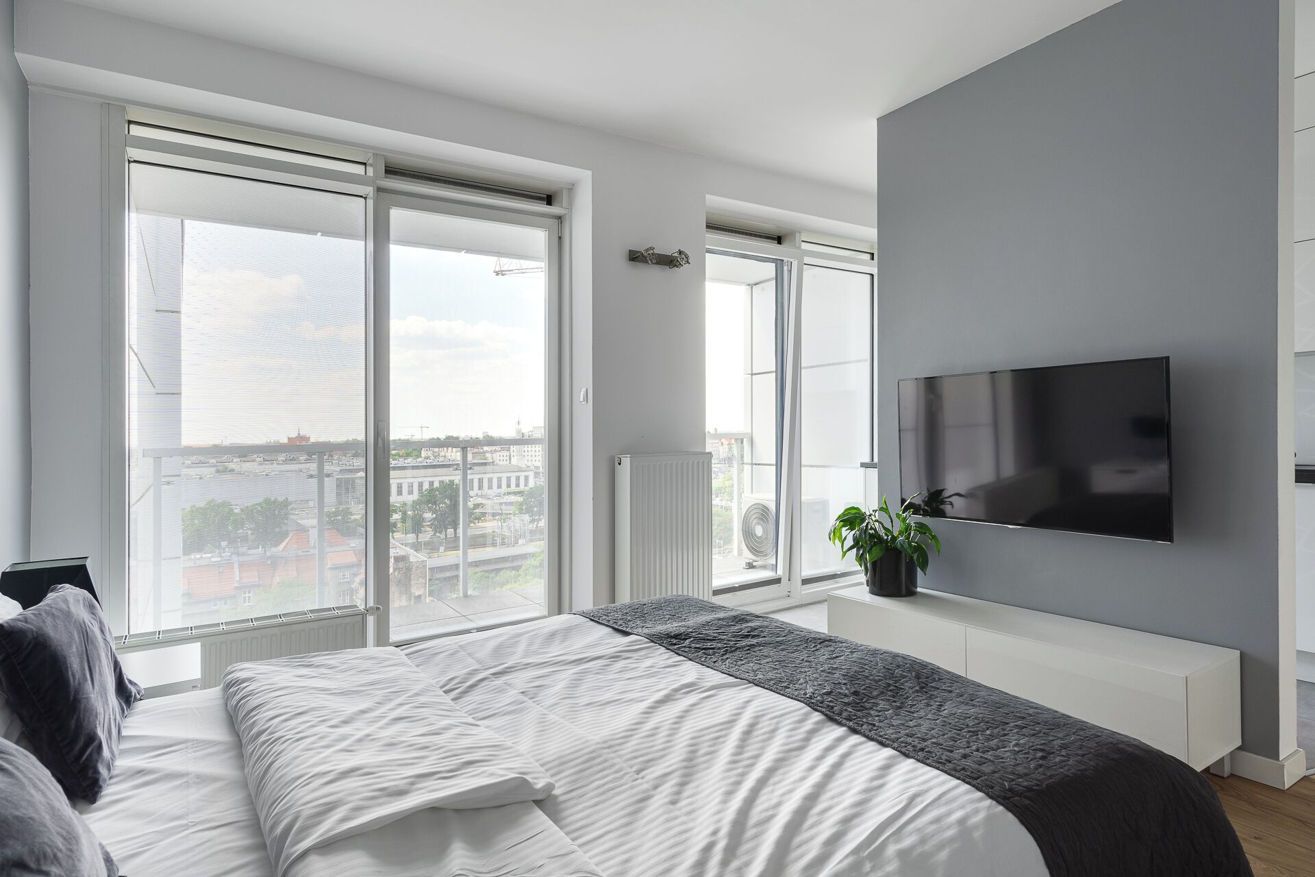 A modern bedroom with a large bed, a TV mounted on the wall, and a view of the city through large windows.