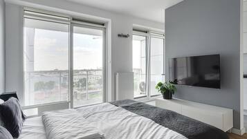 A modern bedroom with a large bed, a TV mounted on the wall, and a view of the city through large windows.
