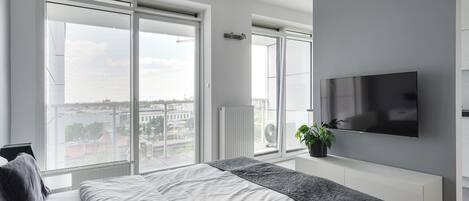 Un dormitorio moderno con una cama grande, un televisor montado en la pared y vistas de la ciudad a través de amplios ventanales.