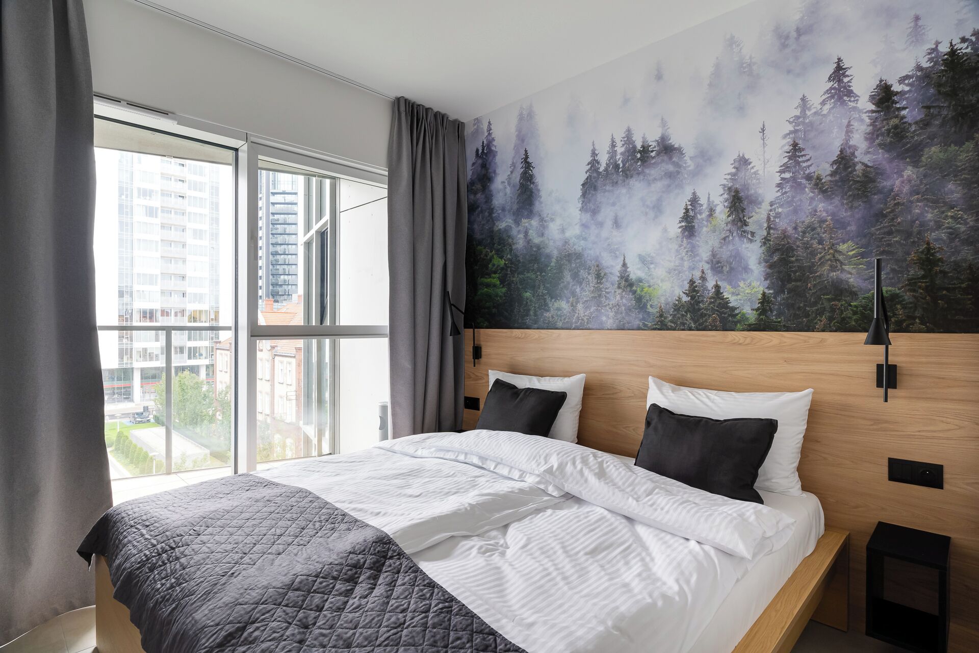 A modern hotel room with a large bed, a view of a cityscape through the window, and a forest-themed wall mural.