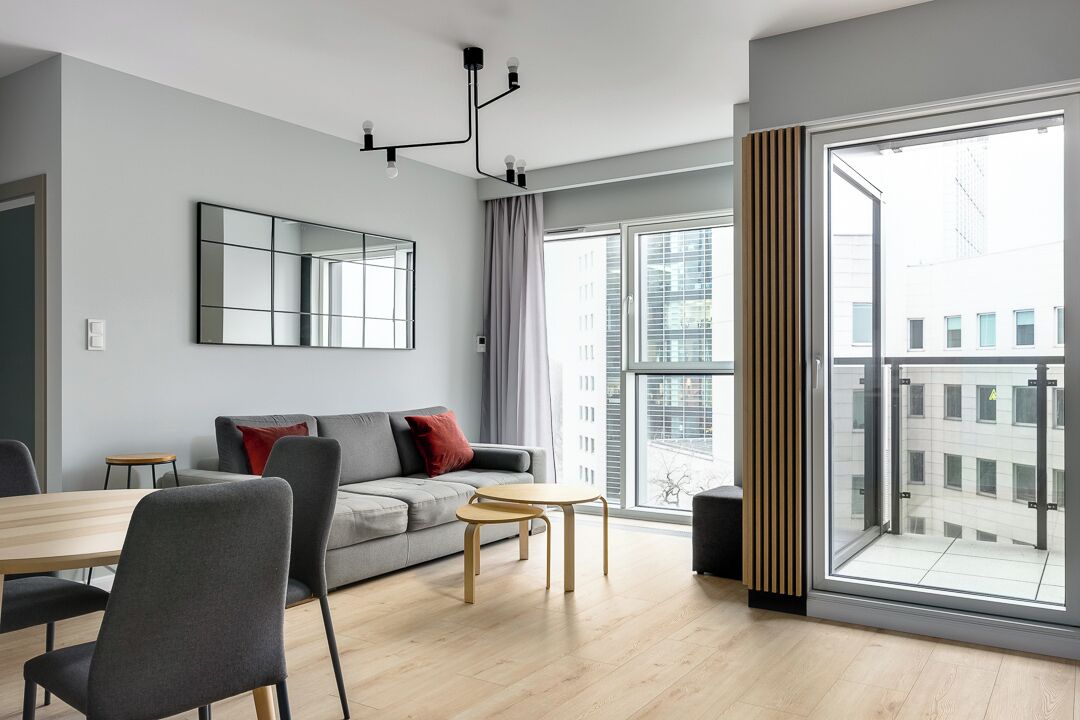 A modern living room with a sofa, a dining table, and chairs. There is a large window with a view of buildings outside.