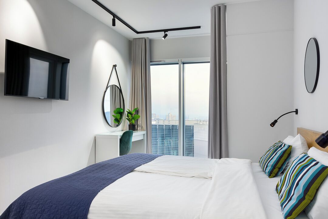 Una habitación de hotel moderna con una cama grande, un televisor montado en la pared, un escritorio con una planta y vistas a la ciudad a través de amplias ventanas.