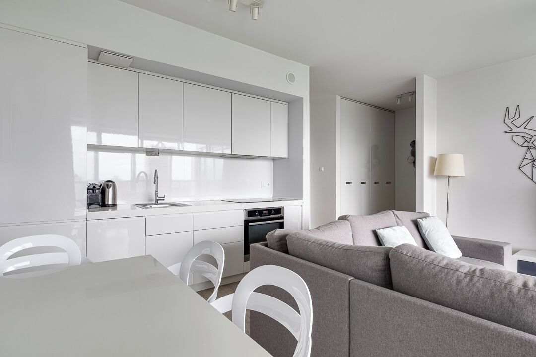 Et moderne køkken med hvide skabe, et spiseområde med et hvidt bord og stole, og en grå sofa i opholdsstuen.