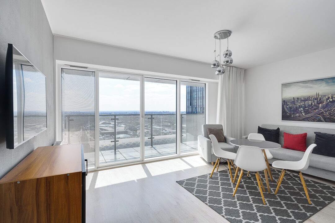 A modern living room with a large sliding glass door leading to a balcony, a flat-screen TV, a sofa with white and red cushions, and a cityscape painting.