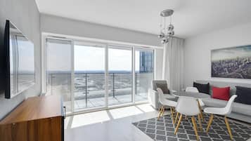 A modern living room with a large sliding glass door leading to a balcony, a flat-screen TV, a sofa with white and red cushions, and a cityscape painting.