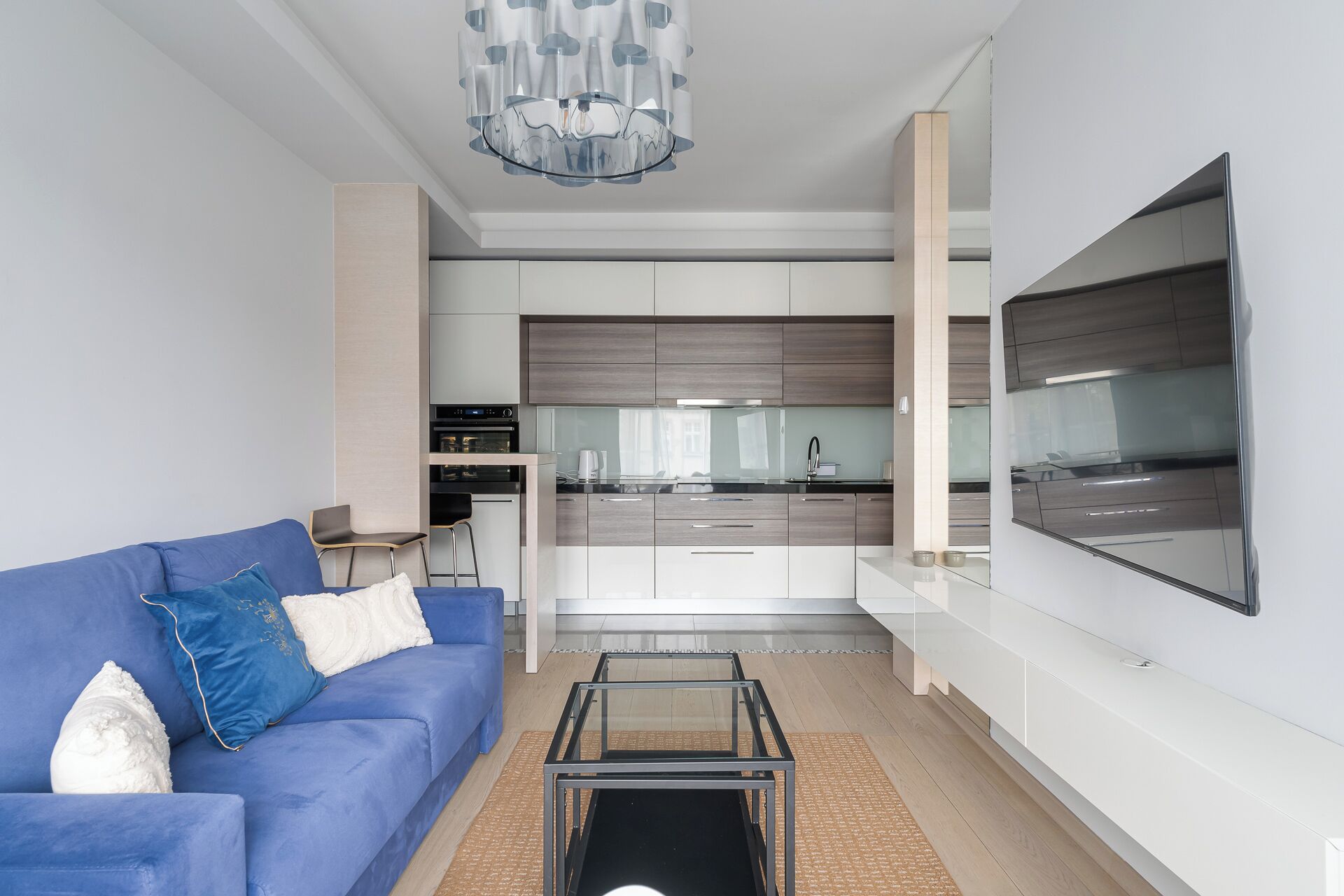A modern living room with a blue sofa, a glass coffee table, and a kitchen in the background.