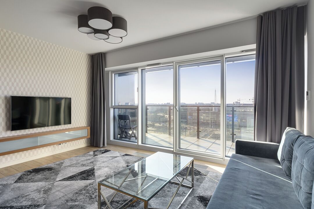 Een moderne woonkamer met een grote televisie, een glazen salontafel, een grijze bank en een stadszicht door grote ramen.