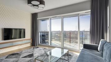 Een moderne woonkamer met een grote televisie, een glazen salontafel, een grijze bank en een stadszicht door grote ramen.