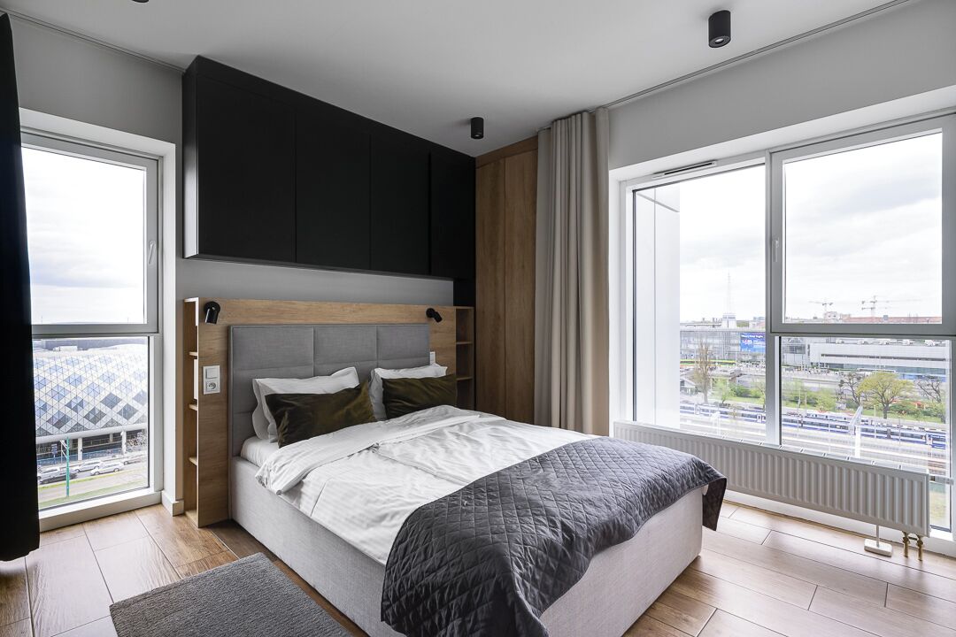 A modern bedroom with a large bed, built-in headboard, and a view of the city through large windows.