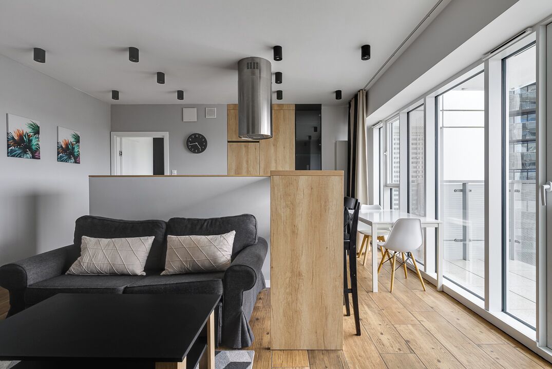 Een moderne woonkamer met een bank, een eettafel en stoelen. Er is een keuken met kasten en een fornuis. De kamer heeft grote ramen die voor natuurlijk licht zorgen.