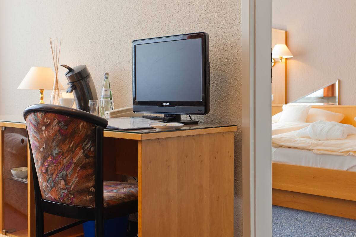 A hotel room with a wooden desk, a computer, a chair, a bed with white bedding, and a lamp.
