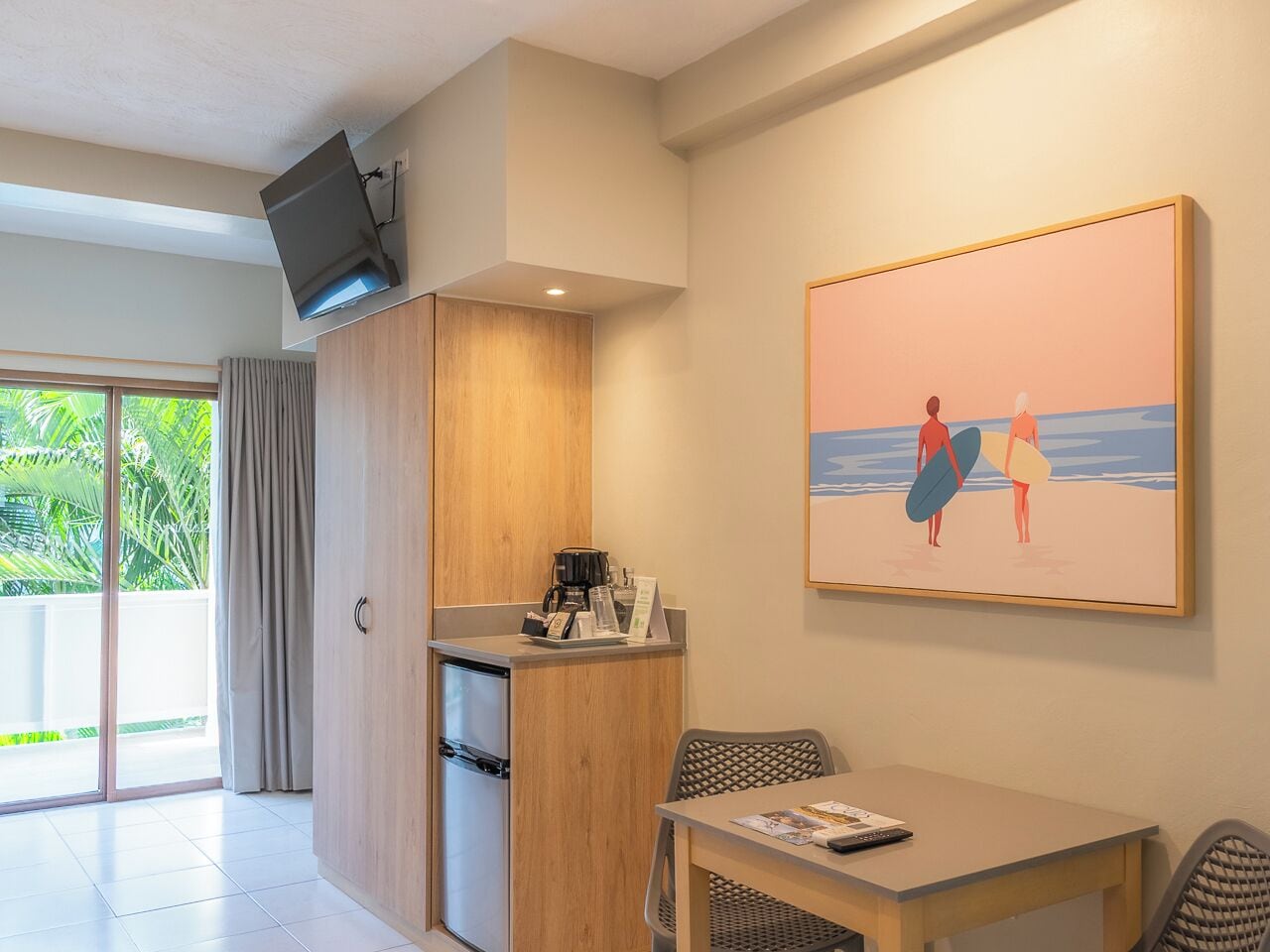 A room with a kitchenette, a dining table, a TV mounted on the wall, and a painting of beachgoers.