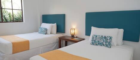 Habitación de hotel con dos camas individuales, una mesita de noche con lámpara y ventana con vistas a palmeras.