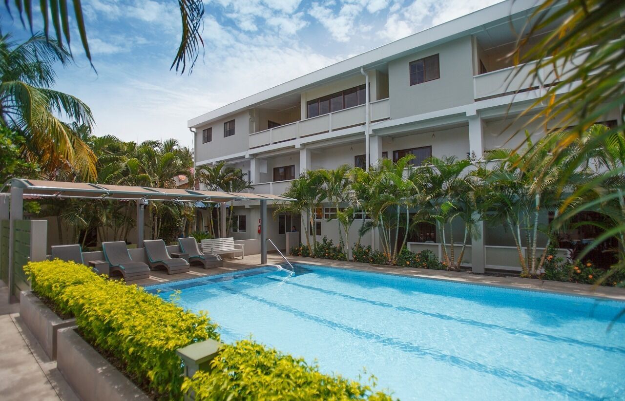 A swimming pool with lounge chairs and a covered area, surrounded by palm trees and a multi-story building.
