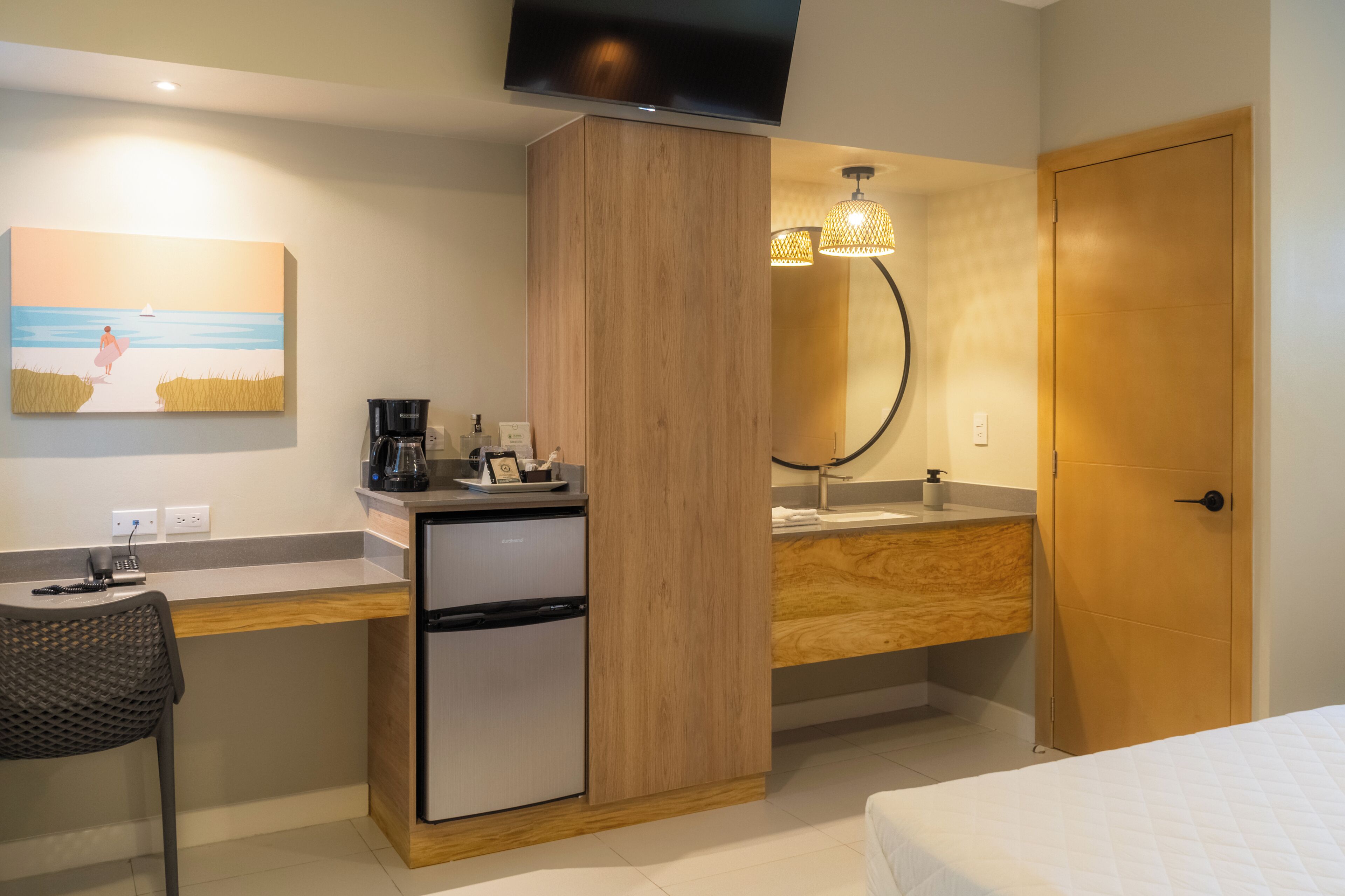 A compact hotel room with a kitchenette, a single bed, a desk with a chair, a TV mounted on the wall, and a painting of a beach scene.