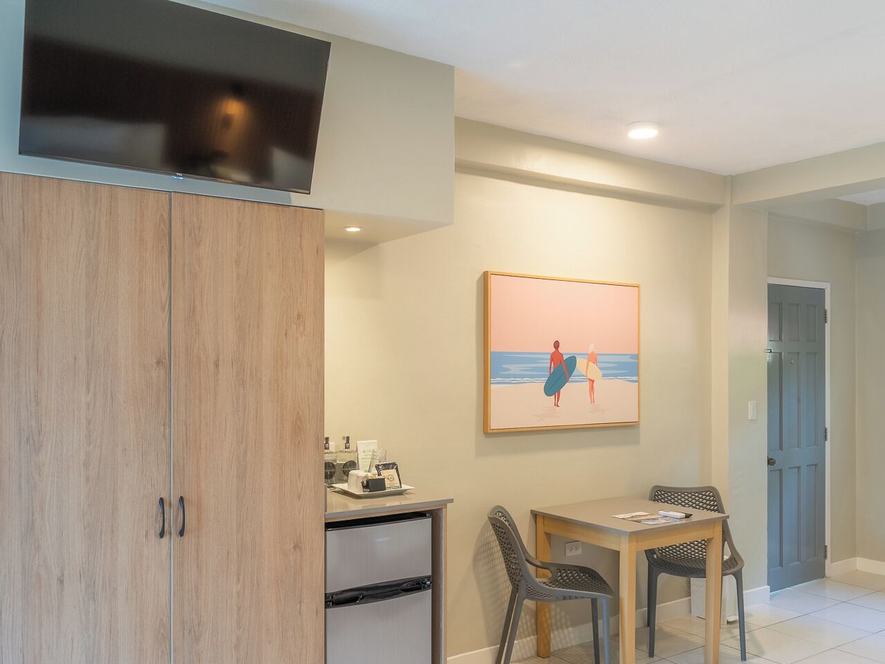 A room with a wooden wardrobe, a desk with chairs, a framed picture on the wall, and a door in the background.