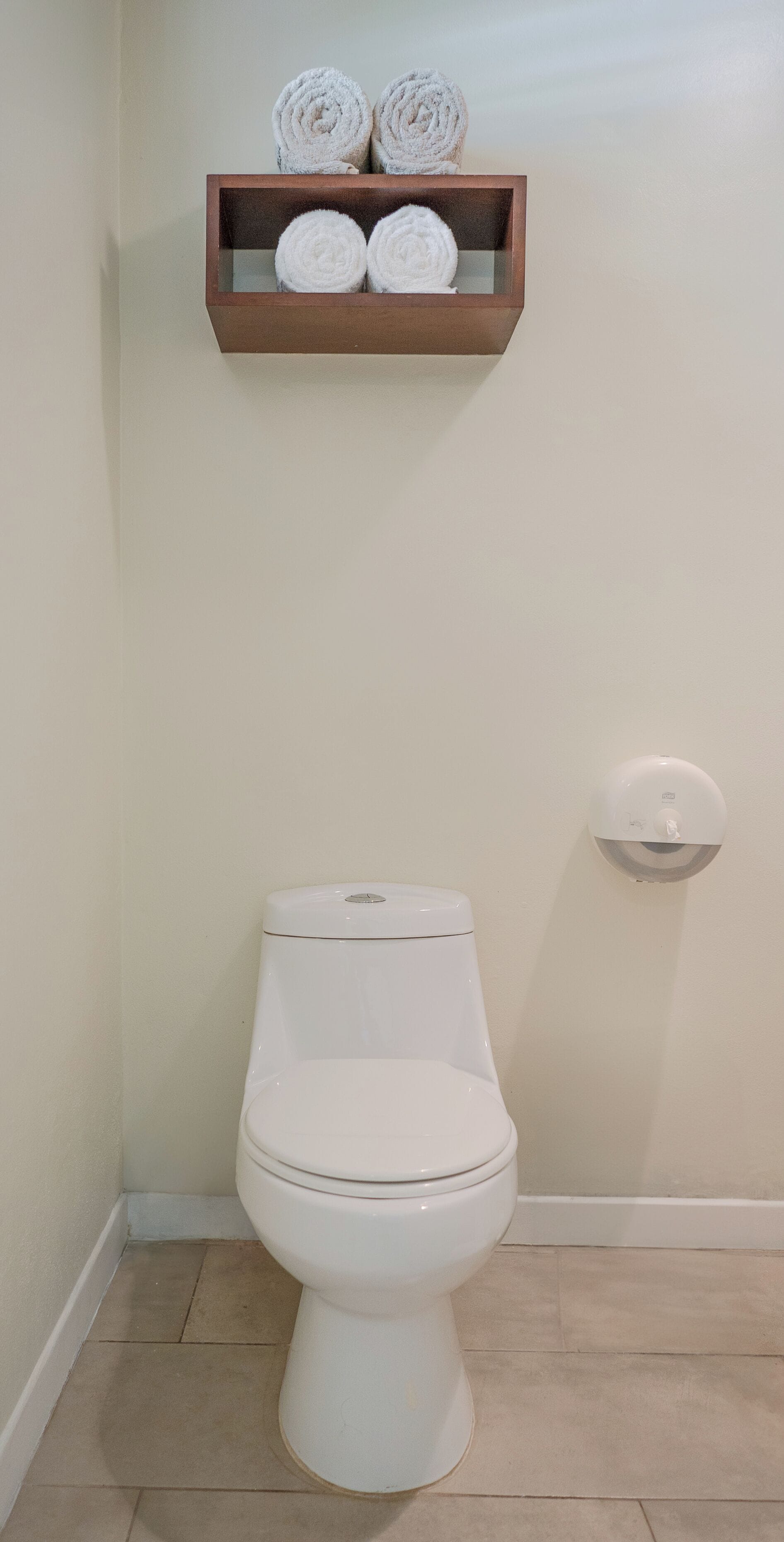 A toilet with a white lid, a wall-mounted toilet paper holder, and a wooden shelf with rolled towels.