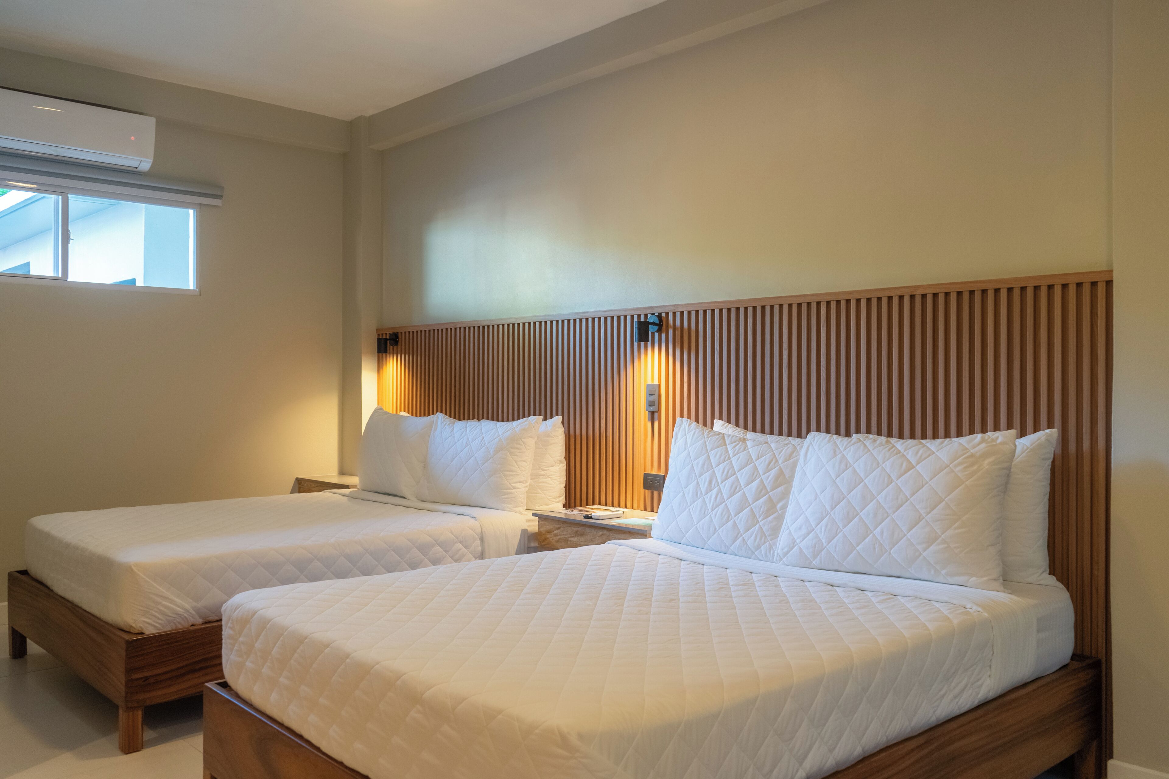 A hotel room with two beds, a window, and an air conditioning unit.