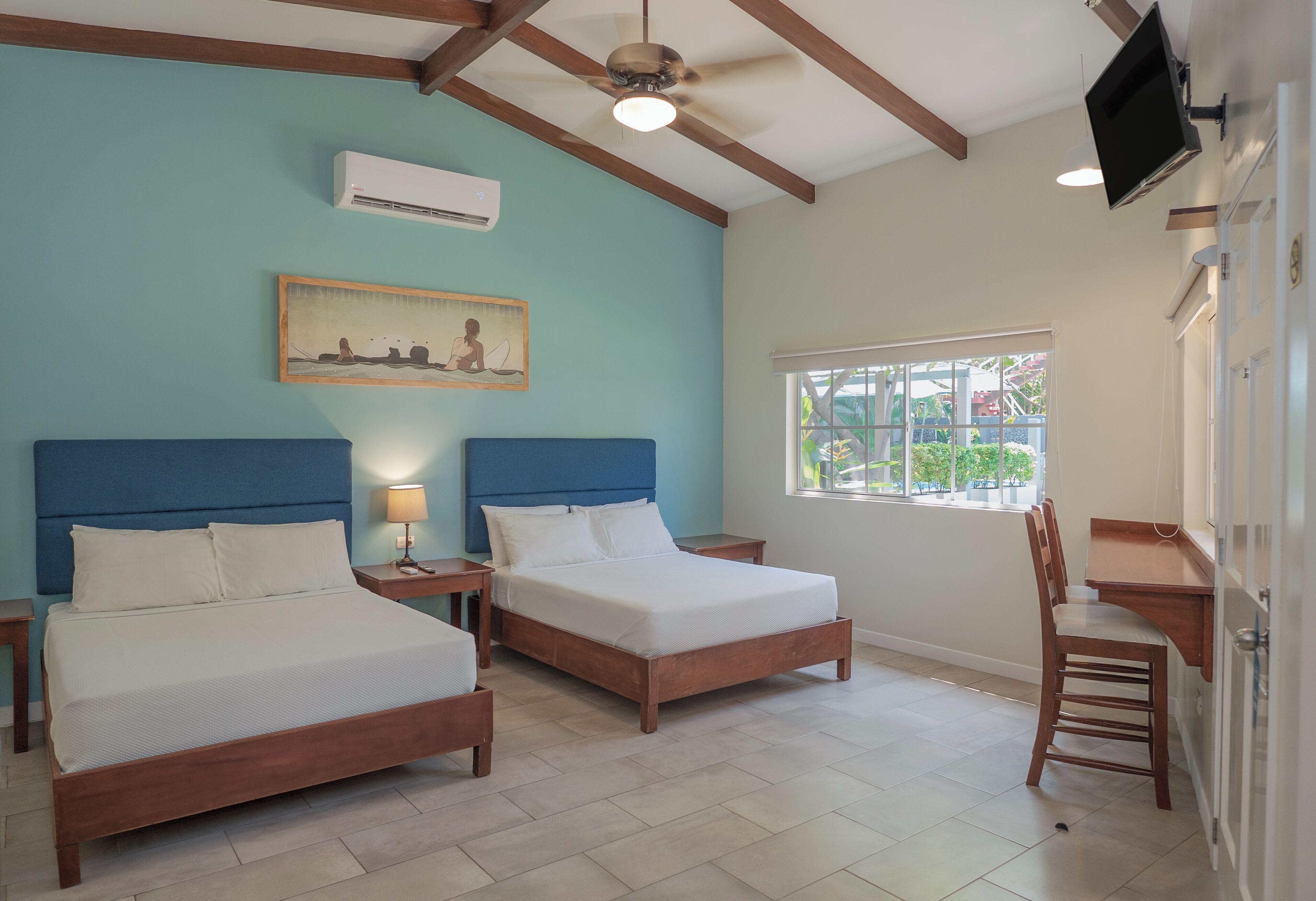 Habitación de hotel con dos camas, ventilador de techo, unidad de aire acondicionado, un cuadro, ventana con vistas y televisor montado en la pared.