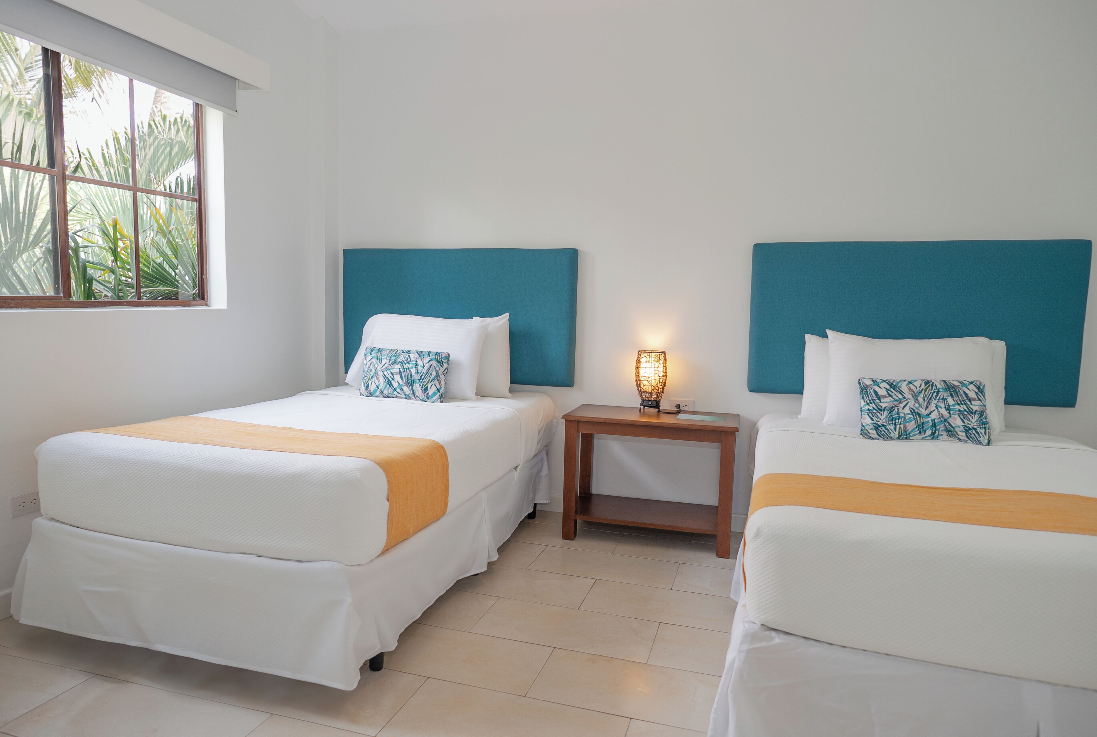 A hotel room with two beds, a nightstand, and a window with a view of palm trees.