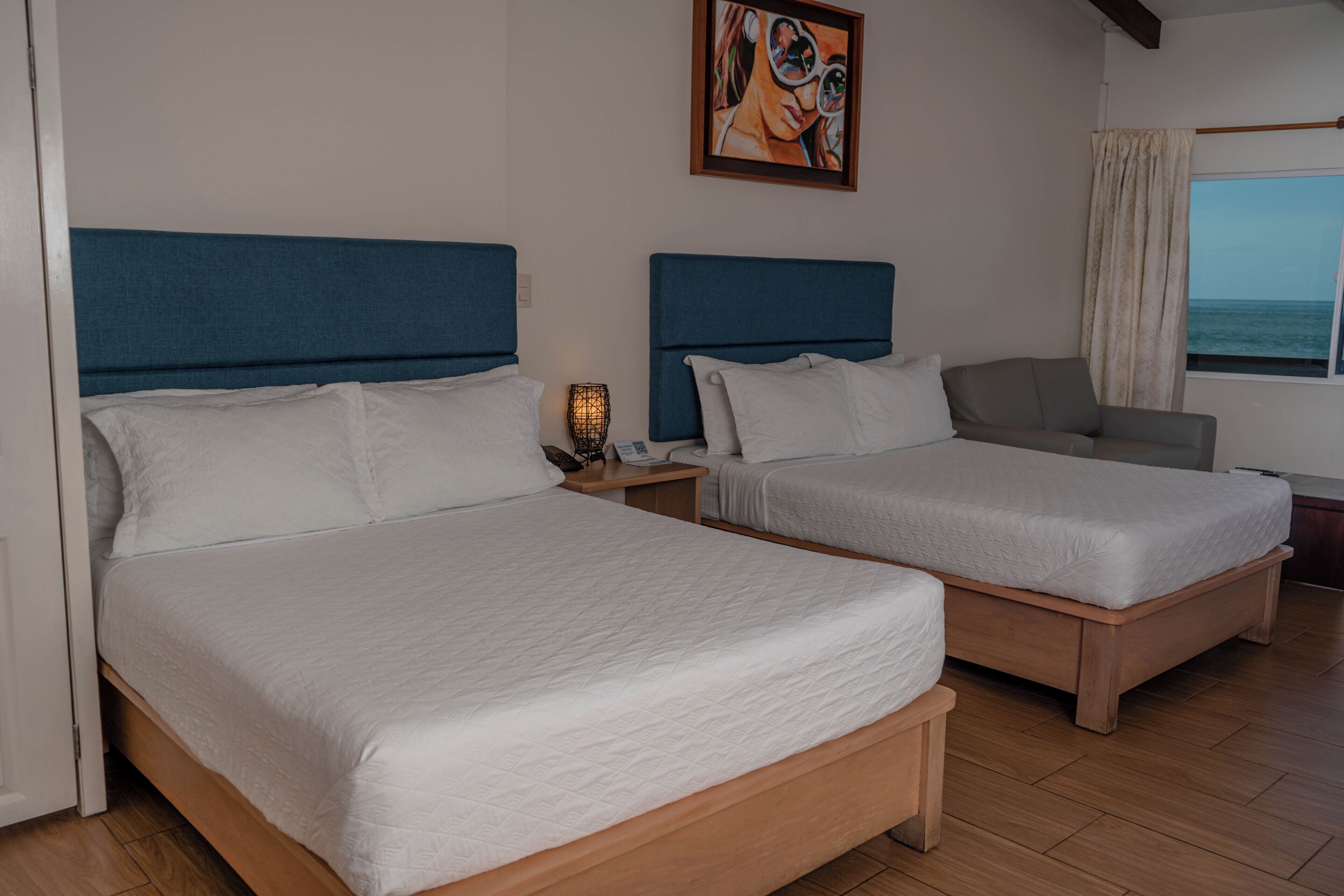 Une chambre d’hôtel avec deux lits, une table de chevet, une chaise, un tableau et une fenêtre donnant sur la mer.