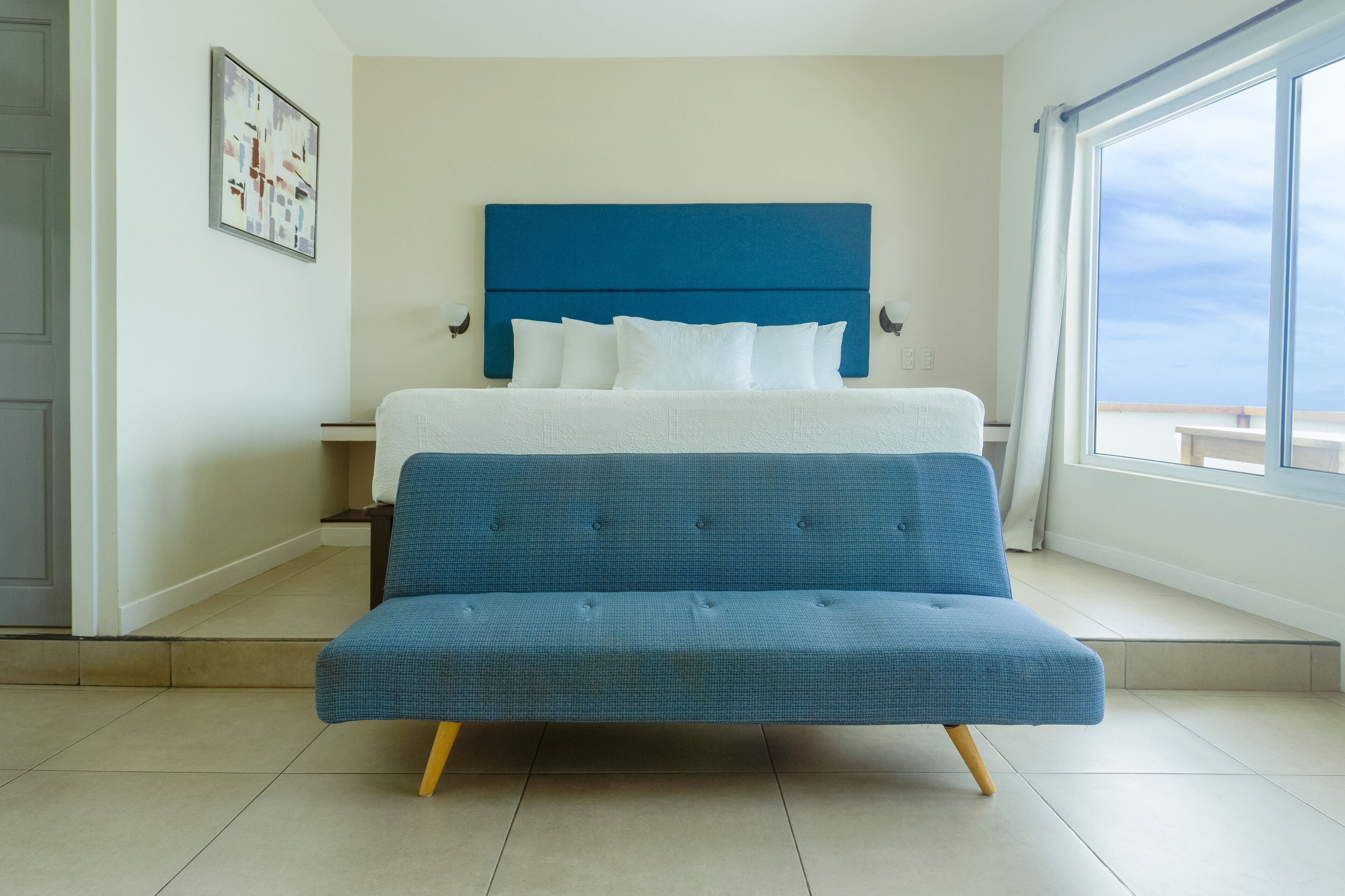Ein Bett mit blauer Kopfteil und weißer Bettwäsche, ein blaues Sofa und ein großes Fenster mit Blick.