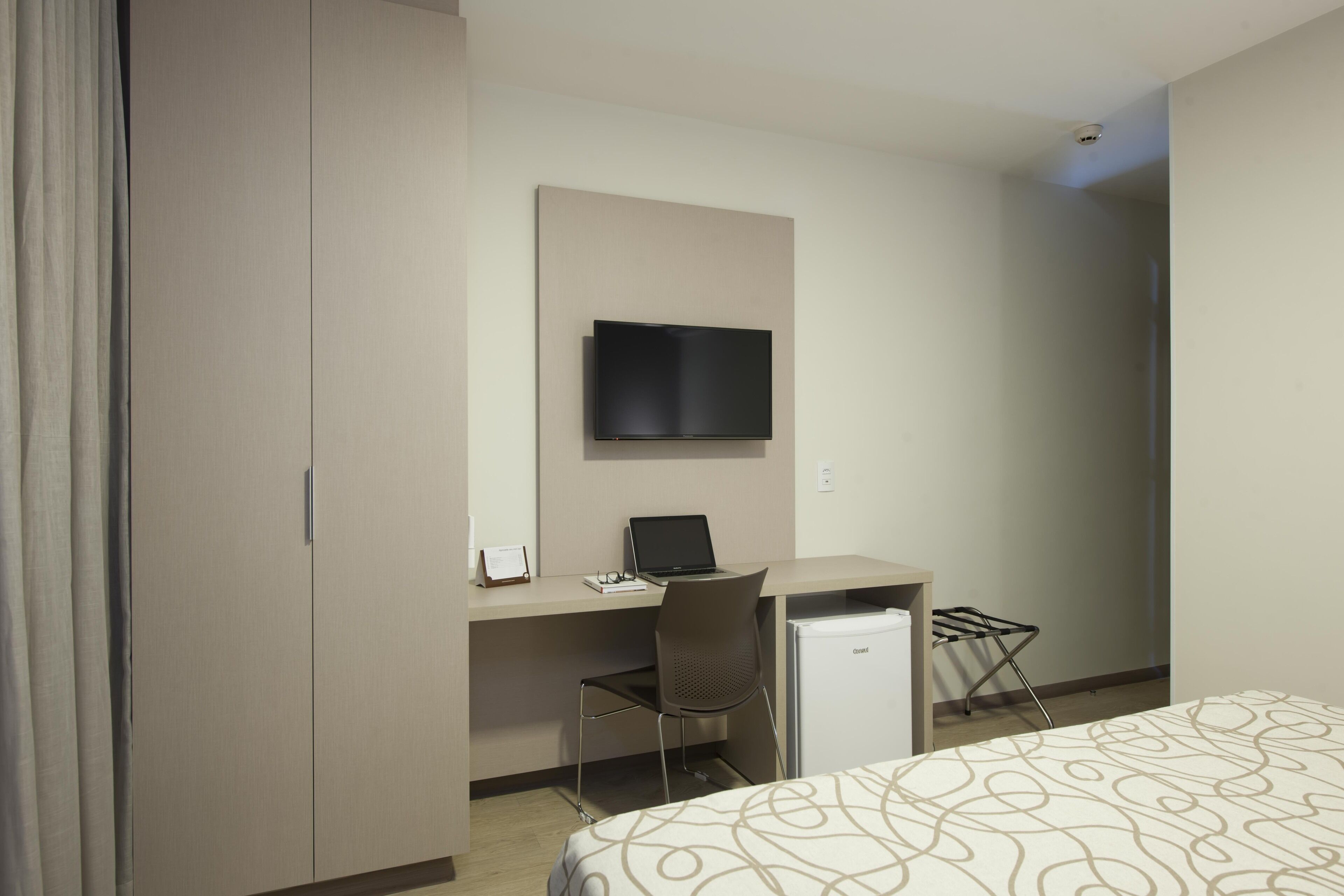 Quarto de hotel com cama, escrivaninha, laptop, televisão e mini-frigorífico.
